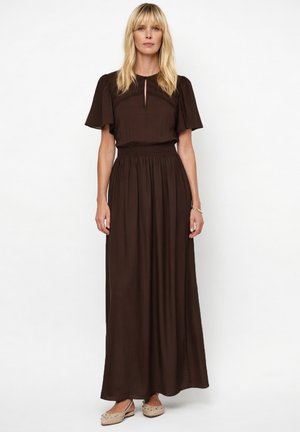 Blonde vrouw die een donkerbruine maxi-jurk met korte mouwen draagt en beige platte schoenen met versieringen, staand tegen een witte achtergrond.