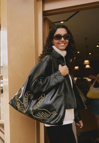 Zwarte leren handtas met gouden stud-zwanelement, glanzende afwerking, oversized vorm, gecombineerd met een zwarte leren jas en een witte coltrui.