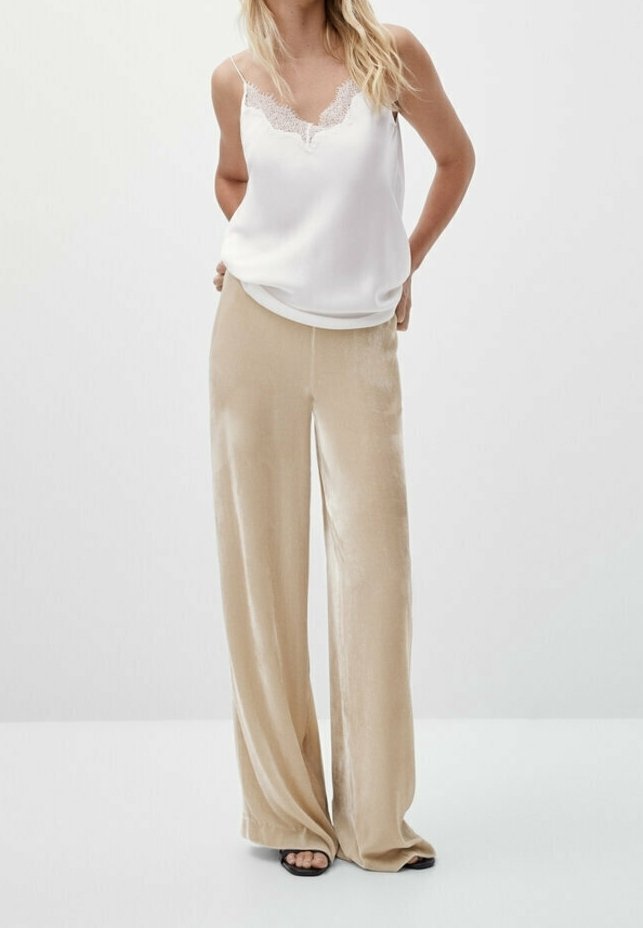 Vit satin cami med spetsdetaljer, kombinerad med vida beige byxor i mjukt tyg. Modellen står med händerna i fickorna.