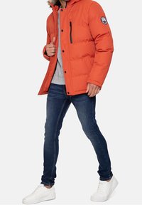 Homme portant une doudoune d'hiver orange vif, un sweat-shirt gris, un jean bleu slim et des baskets blanches, marchant avec une main sur la veste.