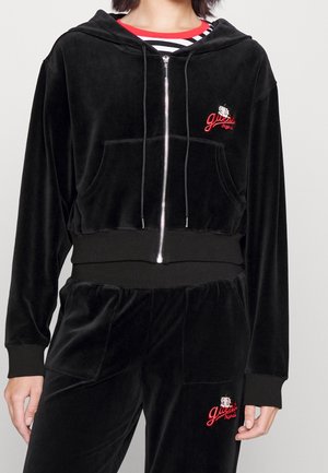 Frau trägt schwarzen Velours-Zip-Hoodie und passende Hose mit roten gestickten Logos, kombiniert mit einem schwarz-weiß gestreiften Shirt mit rotem Halsausschnitt.