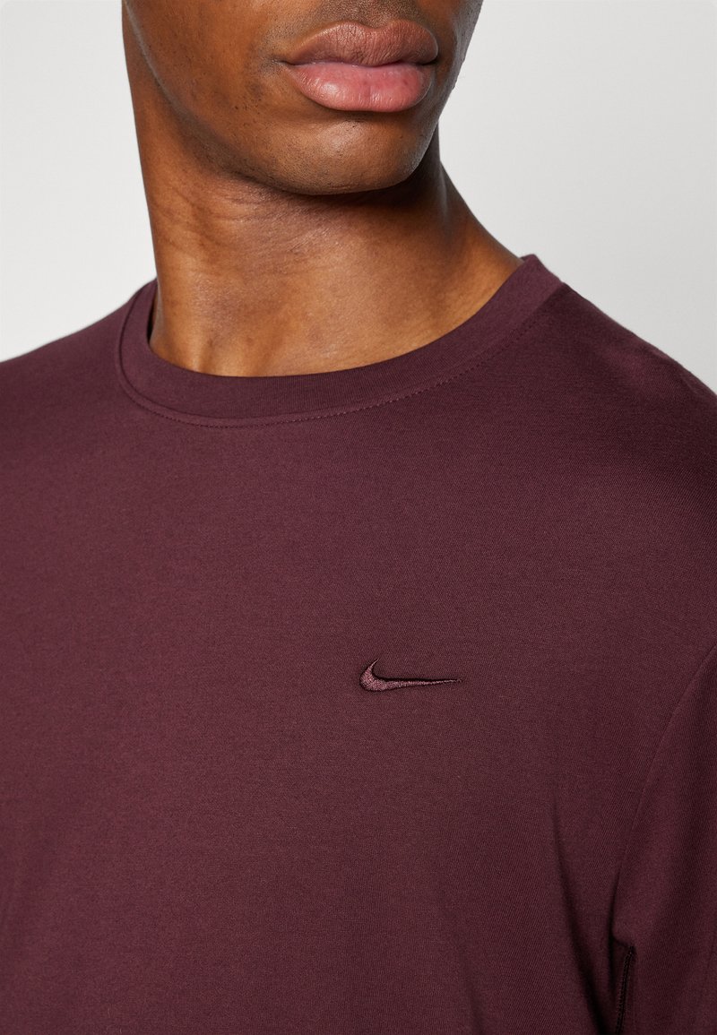 Burgundowa koszulka Nike z okrągłym dekoltem, krótkimi rękawami i subtelnym haftem logo na klatce piersiowej. Gładka bawełniana struktura.