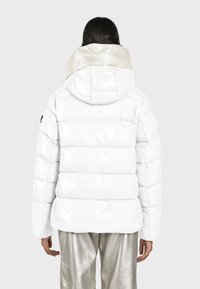 Witte pufferjack met een glanzende afwerking, voorzien van een contrasterende beige capuchon. Paneelontwerp met zichtbare stiksels. Draagt over metalen zilveren broeken.