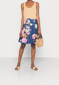 Stickad beige linne, marinblå knälång kjol med blommönster, vävd handväska och nubuck tofflor. Har livfulla rosa och orange blommor.