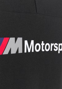 Černá tkanina s výrazným šedým písmenem "M" a textem "Motorsport"; na levé straně jsou viditelné červené a modré akcenty. Hladká textura.
