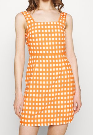 Frau trägt ärmelloses orange-weiß kariertes Kleid mit quadratischem Ausschnitt, steht vor einem einfarbigen Hintergrund.