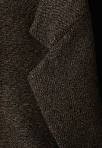Tessuto di lana marrone con una superficie texturizzata, caratterizzato da un motivo sottile. Il materiale si drappeggia in modo elegante con una trama leggermente irregolare.
