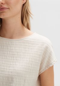 Lähikuva naisesta, jolla on päällä tekstuuriltaan hieman valkoinen, lyhythihainen paita, jossa on pyöreä kaula-aukko, vaaleataustaista yksinkertaista taustaa vasten.