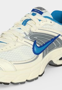 Λευκό και μπλε αθλητικό παπούτσι Nike Moto 2K με χοντρή σόλα, λεπτομέρειες από διχτυωτό ύφασμα, γκρι αποχρώσεις και μπλε λογότυπο Nike στο πλάι.