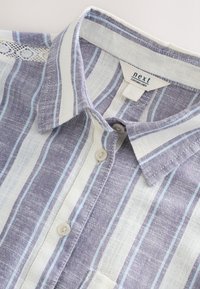 Chemise en lin avec un col, présentant des rayures violettes claires et blanches, un détail décoratif en dentelle et une fermeture par boutons.