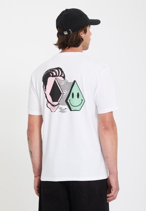 Homme portant un t-shirt blanc avec un motif abstrait coloré de visage souriant au dos, une casquette noire et un pantalon noir, debout devant un fond blanc.