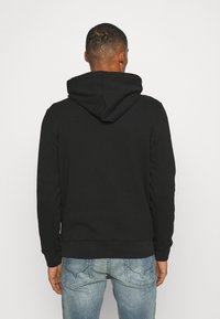 Uomo vestito con un hoodie nero e jeans blu, rivolto di spalle contro uno sfondo bianco uniforme.