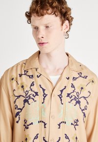 Beige overhemd met korte mouwen en kraag, voorzien van blauwe en groene bloemembroidery en knoopsluiting aan de voorkant. Zachte textuur.