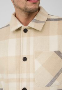 Close-up van een man in een beige geruite overhemd met zwart knopen en een borstzakje, waarbij een deel van zijn gezicht en nek zichtbaar is.