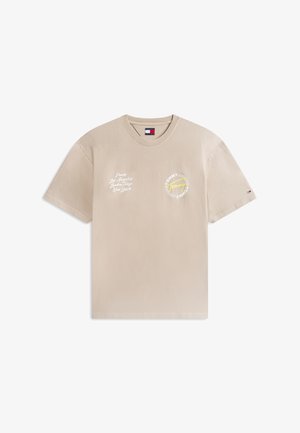 Šviesiai smėlio spalvos trumpomis rankovėmis marškinėliai su baltomis miesto pavadinimų užrašais kairėje krūtinės pusėje ir apvaliu gelsvos ir baltos spalvų Tommy Jeans logotipu dešinėje krūtinės pusėje.