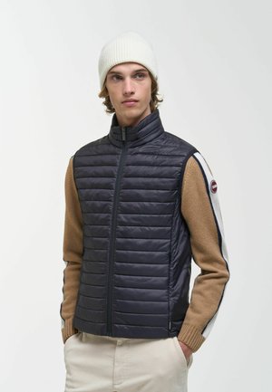 Junger Mann, der eine weiße Strickmütze, eine marineblaue gesteppte Weste, einen beigen Pullover mit weißen und marineblauen Streifen und beige Hosen trägt, steht mit den Händen in den Taschen.