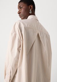 Camicia con bottoni, a righe verticali beige e bianche, con dettaglio sul retro e maniche lunghe con bottoni ai polsi. Tessuto di texture liscia.