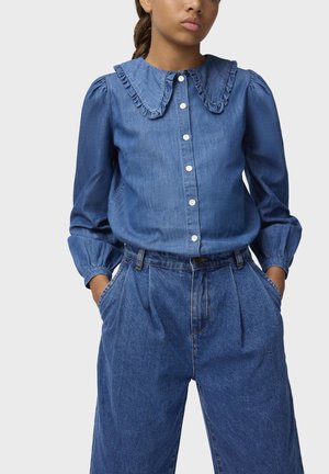 Ung person iført blå denim knappet bluse med flæsekant og højttaljede blå denim bukser, hænderne i lommerne.