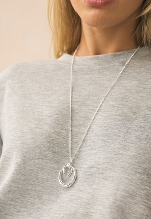 Woman wearing a silver chain necklace with two linked oval pendants over a light grey crew-neck top.