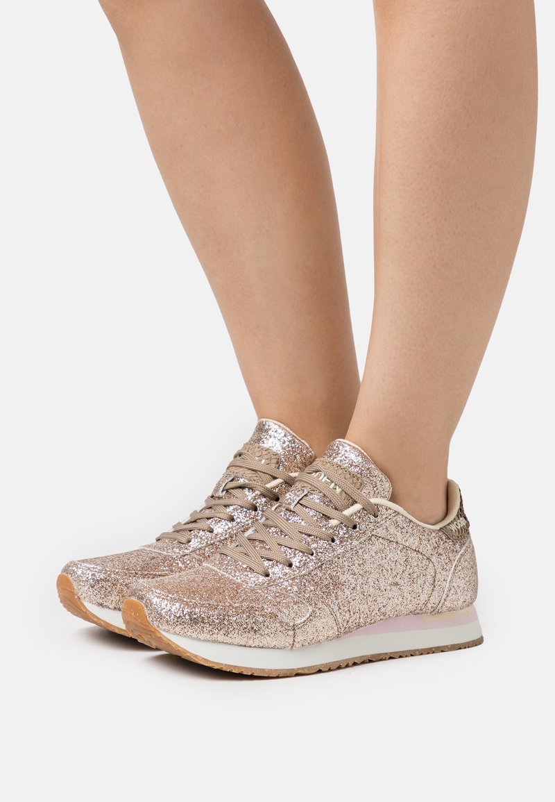 Jambes portant des baskets scintillantes rose gold avec des lacets beiges et des semelles marron clair, se tenant sur un fond blanc.