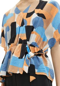 Blusa colorata e stampata con forme astratte in blu, arancione e nero. Tessuto morbido con vita arricciata e dettaglio con cravatta. Scollatura a V.