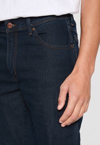Nahaufnahme einer Person, die dunkelblaue Denim-Jeans und ein weißes Hemd trägt. Zu sehen ist die vordere Tasche und die rechte Hand, die neben dem Bein ruht.
