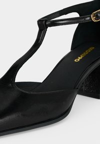 Chaussure à talon en cuir noir avec bride en T, bout pointu, talon bloc et boucle dorée.
