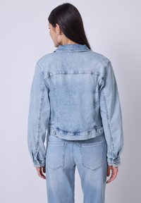 Donna con lunghi capelli scuri che indossa grandi orecchini a cerchio, una giacca di denim azzurro chiaro e jeans abbinati, vista di spalle su uno sfondo semplice.