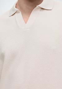 Personne portant une chemise texturée couleur crème à col avec un col en V ouvert. Seuls le cou et le haut de la poitrine sont visibles.