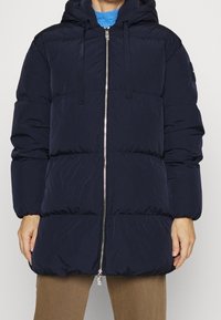 Námořnická péřová bunda s kapucí, která má plný zip vpředu, elastické manžety a vodorovné prošívané části pro zvýšené teplo a pohodlí.