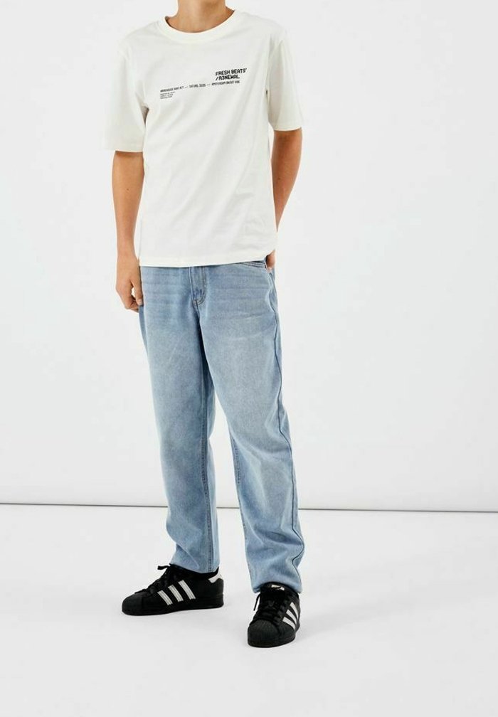 Camisa blanca de manga corta con texto negro; jeans sueltos de color azul claro; zapatos negros con tres rayas blancas. Un atuendo simple y casual.