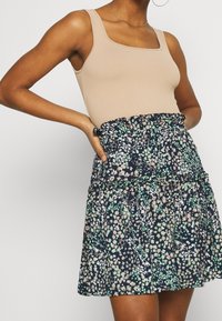 Tanktop in lichtbeige, gecombineerd met een marineblauwe rok met bloemenprint. De rok heeft een geplooide taille en gelaagde ruches met pastelaccenten.