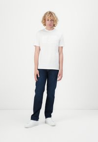White cotton t-shirt featuring embossed "GUESS" logo, paired with dark blue jeans and white sneakers. Model standing on a white background.