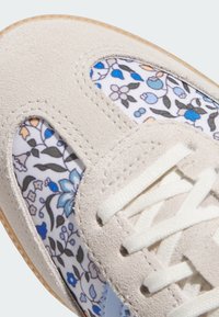 Sneaker mit einem hellgrauen Wildleder-Obermaterial, weißen floralen Textilpartien mit blauen und orangefarbenen Akzenten sowie weißen Schnürsenkeln. Gummisohle.