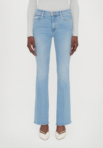 Jeans évasés bleu clair à taille haute, avec poches avant et décoloration subtile, associés à des talons aiguilles blancs.