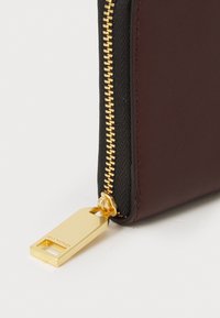 Portafoglio bordeaux e nero con zip dorata e un cinturino rettangolare, che presenta una texture in pelle liscia e linee pulite.
