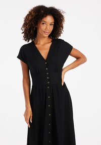 Robe noire à boutons avec un décolleté en V, des manches courtes, une taille cintrée et une jupe fluide. Tissu texturé avec des boutons visibles sur le devant.
