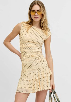 Kvinde iført en gul prikket ruched kjole, ravfarvede solbriller, der holder en striped taskestrop mod en ensfarvet baggrund.