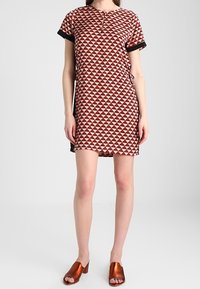 Kurzes ärmelloses Kleid mit einem rot-weißen Dreiecks-Muster, schwarzem Besatz und einem verstellbaren Band in der Taille. Getragen mit bronzefarbenen Slippern.