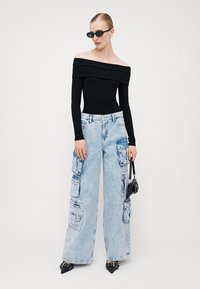 Melnā off-the-shoulder ribota blūze, gaiši zili platie kabatās bikses ar vairākiem kabatām un rāvējslēdzējiem, kā arī melni norādītās kurpes ar sprādzēm.