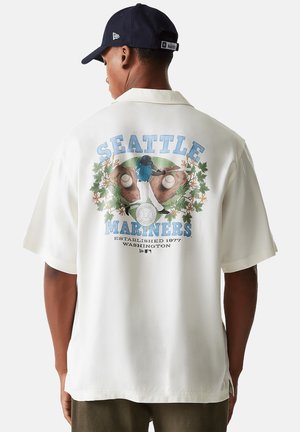 Mann trägt weißes Seattle Mariners Shirt mit Grafik eines Baseballspielers und Handschuhs auf dem Rücken und trägt eine dunkelblaue Mütze, schaut weg.