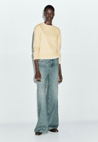 Light yellow sweater with long sleeves, paired with light blue flared jeans and brown shoes. Model stands on a neutral background.