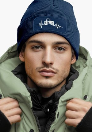 Navy knit beanie with a textured design and a white tractor graphic on a rectangular patch; worn with a green puffer jacket and black layers.