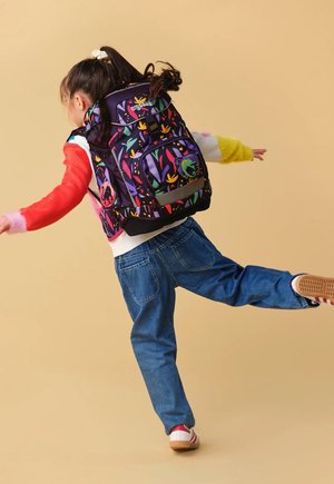 Kind in buntem Pullover und Jeans balanciert auf einem Bein mit großem gemustertem Rucksack vor beigem Hintergrund