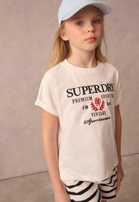 Bílé tričko s krátkým rukávem s potiskem "SUPERDRY" v námořnické modři a červené barvě, spárované s černo-bílými proužkovanými šortkami. Lehký bavlněný materiál.