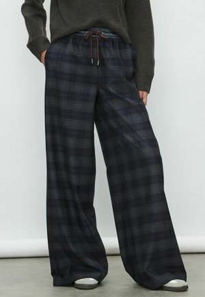 Persona che indossa pantaloni larghi a quadri scuri con vita elastica e cordoncino marrone, abbinati a un maglione scuro e sneakers bianche.