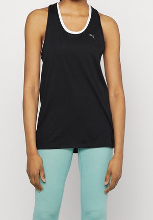 Femme portant un débardeur noir sans manches Puma sur un soutien-gorge de sport blanc et un legging bleu clair, debout devant un fond uni.