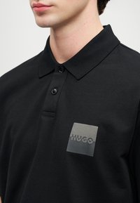Man med svart kortärmad polo med två knappar och en fyrkantig grå logotyp med texten "HUGO" på bröstet.
