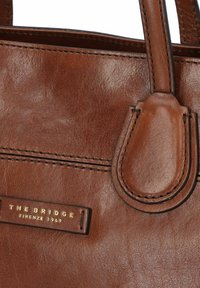 Borsa in pelle marrone con una texture liscia, cuciture visibili e un'etichetta logo dorata che recita "THE BRIDGE, FIRENZE 1969."