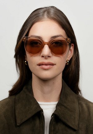 Gafas de sol marrones de gran tamaño con montura marrón clara, con forma geométrica y un sutil detalle de logo, llevadas con una chaqueta de ante oscura.